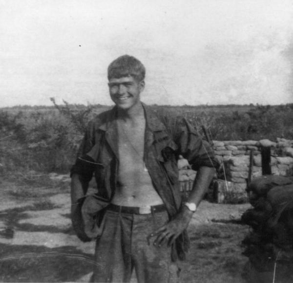 Private First Class Fred Walter Jansonius black and white photo, he is standing outdoors in what appears to be a military encampment. He’s wearing fatigue‑style trousers and an unbuttoned shirt 