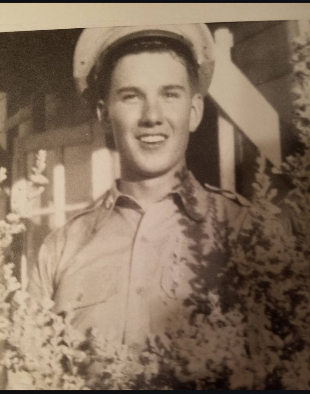 Vintage photograph of Raymond D. Ledoux in a military-style uniform and cap standing outdoors near foliage, with a building structure in the background.