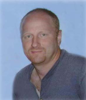 A head-and-shoulders portrait of Kenneth Hendrickson with a shaved head and a light goatee, wearing a grey v-neck shirt.