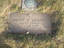 The photograph shows a flat, rectangular military grave marker for Robert C Hausken set into the ground and surrounded by grass.