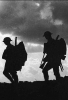 Silhouette of two soldiers walking with helmets, rifles, and heavy packs against a cloudy sky.